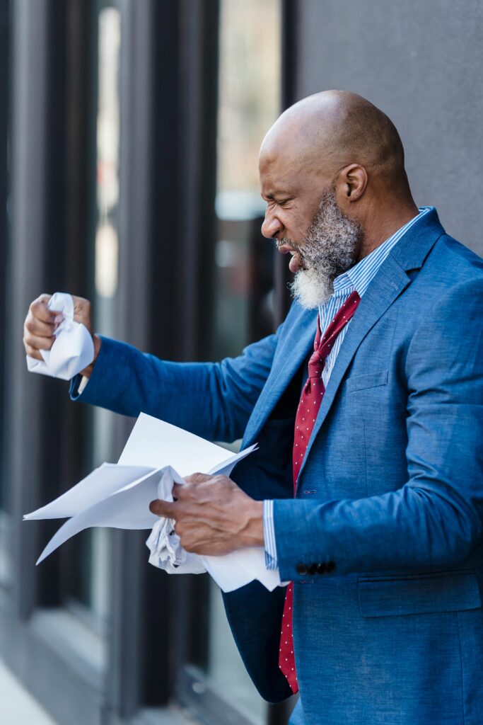현재 이미지: Side view of furious African American male entrepreneur in formal wear with documents in hands standing on street near building
